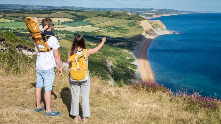 A woman and a man with a baby carrier looking out over the Jurassic Coast from Golden Cap in Dorset, on a bright summer's day